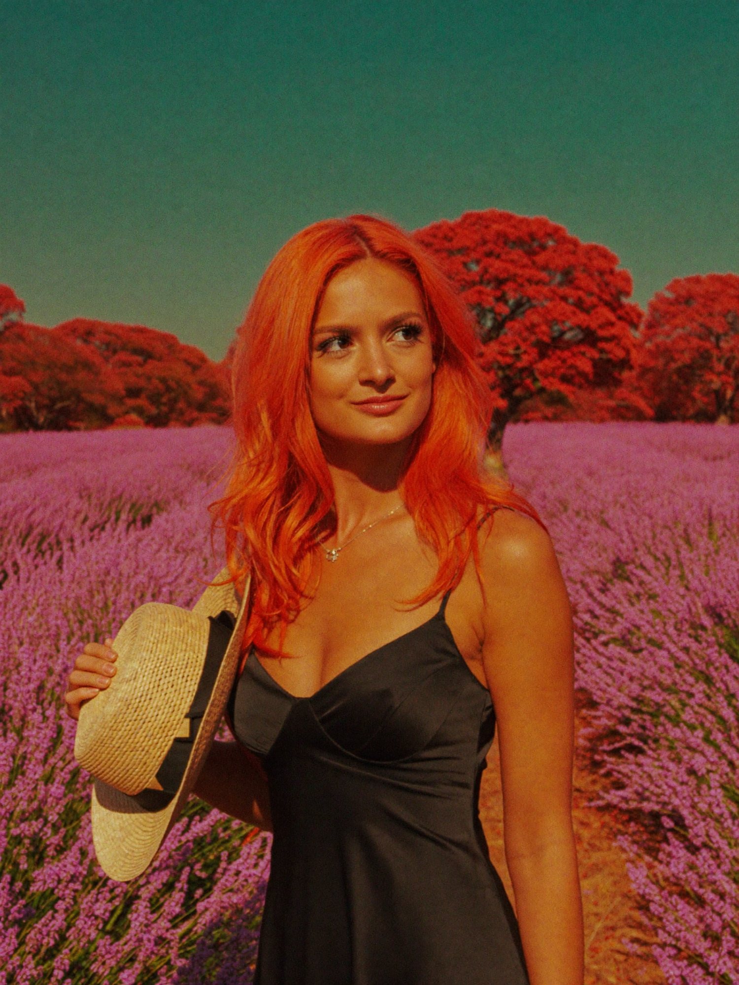 Orange-haired Carla in a black slip holding a straw hat in a lavender field under red trees.