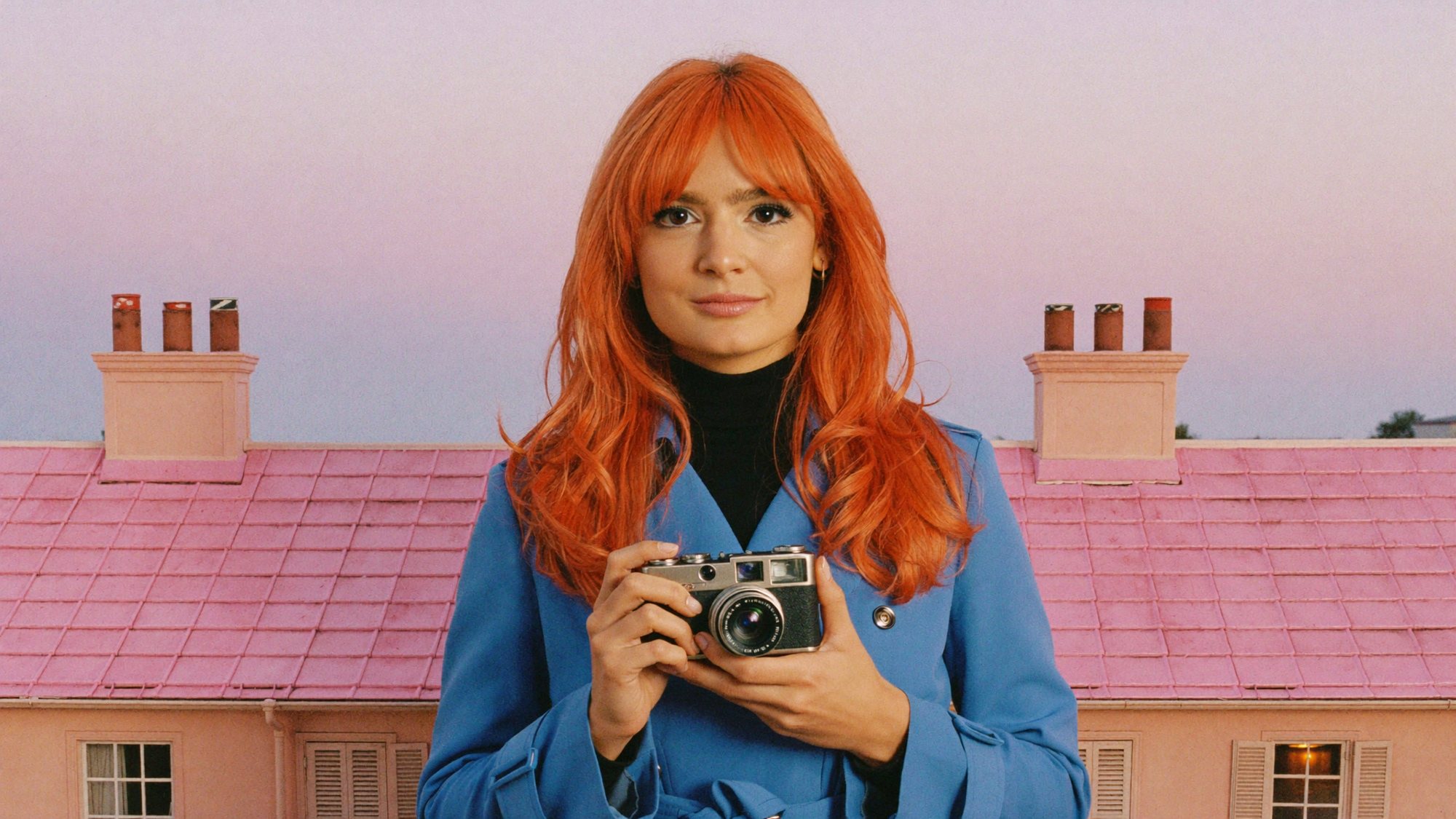 Orange-haired Carla in a blue coat holding a camera on a pink rooftop at dusk.