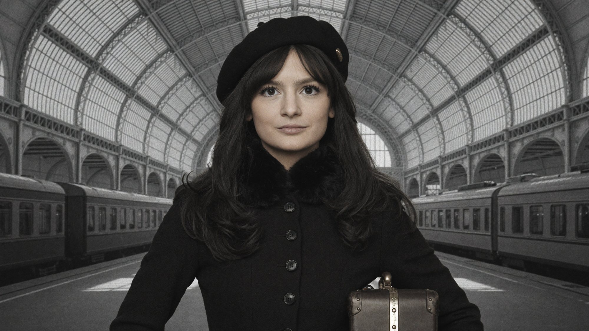 Carla in a black beret and coat with a small suitcase at a railway station, in black and white.