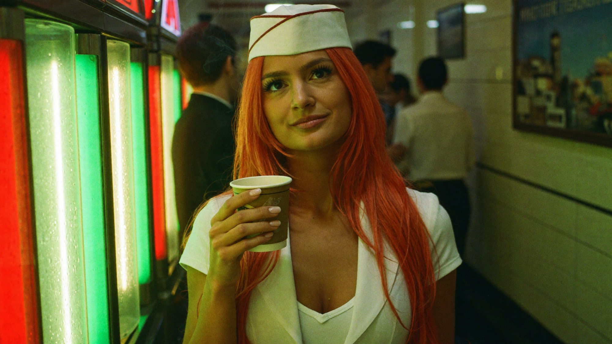 Orange-haired Carla in a white cap holding a paper cup by a neon jukebox in a narrow corridor.