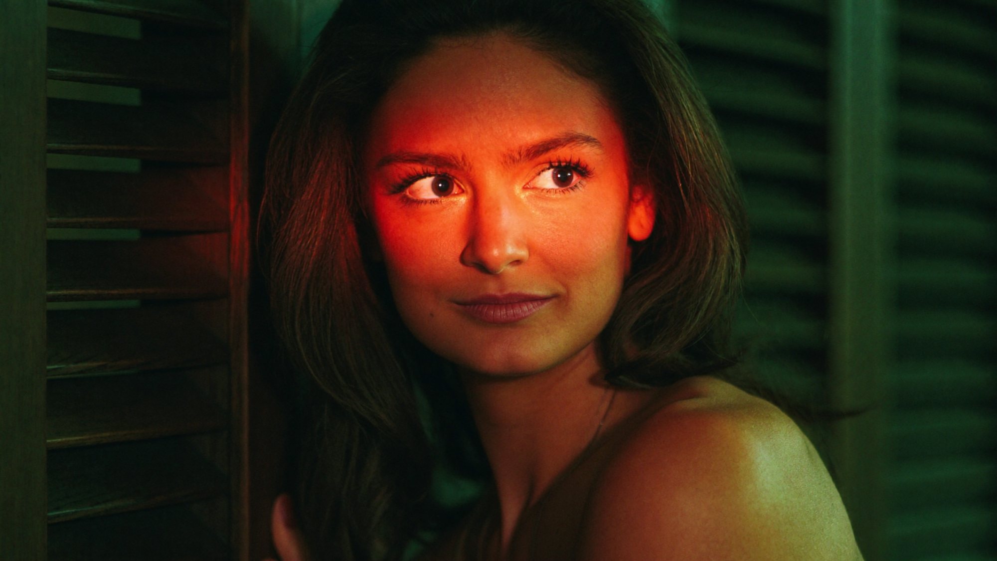 Carla's face lit red through green shutters as she glances sideways.