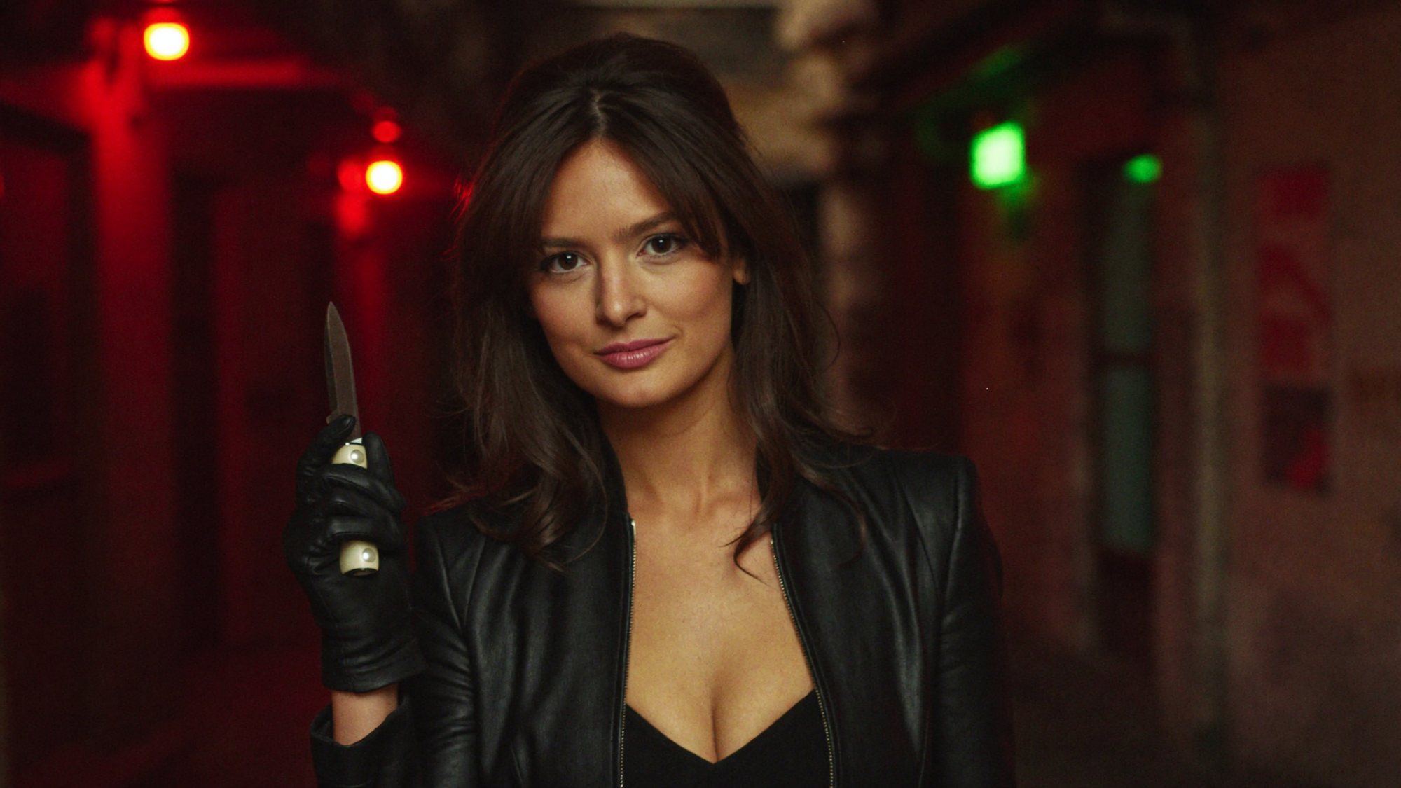 Carla in a black leather jacket holding a knife in a black-gloved hand in a red-lit corridor.