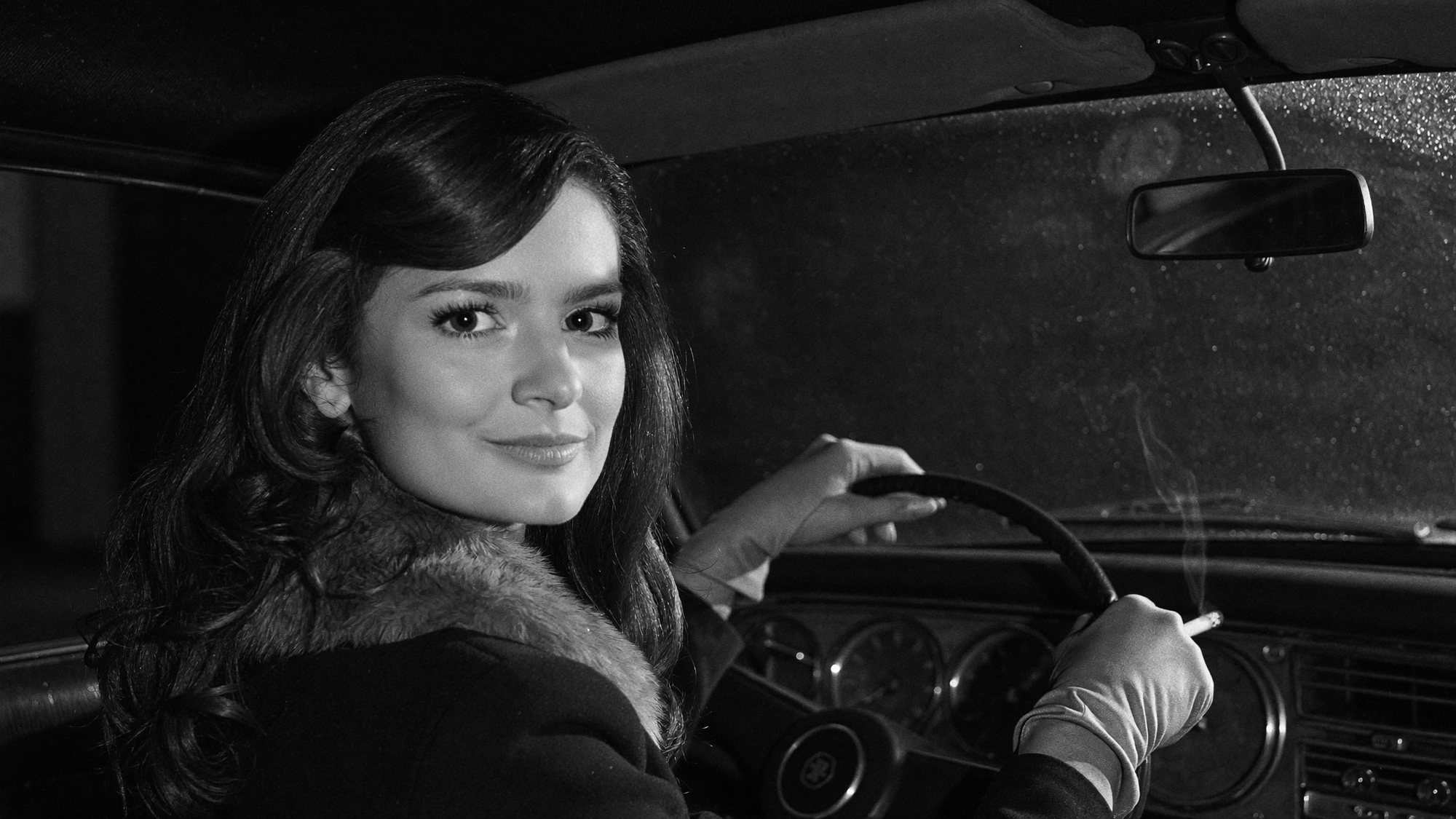 Carla in gloves and a fur collar at the wheel of a car in driving rain. Black and white.