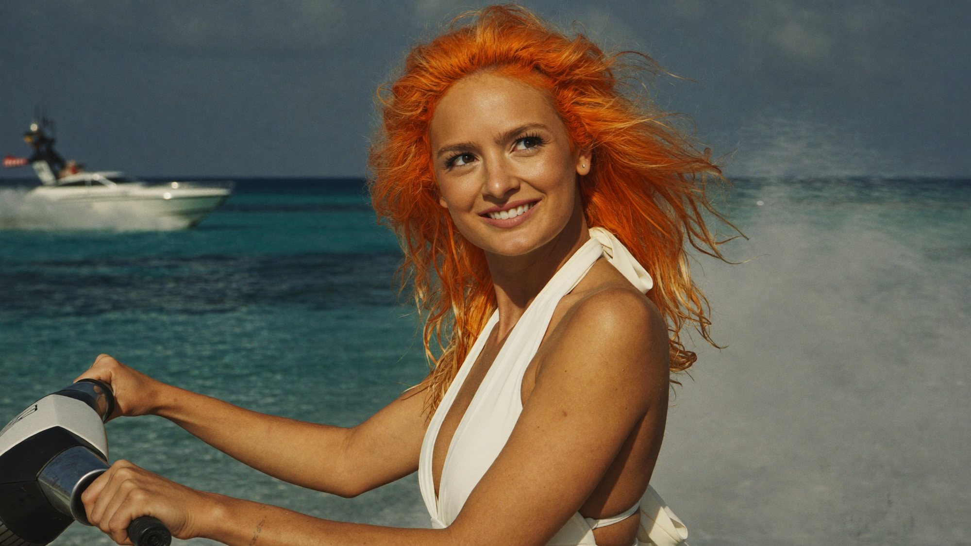 Orange-haired Carla in a white swimsuit riding a jet ski in a chase across turquoise water.