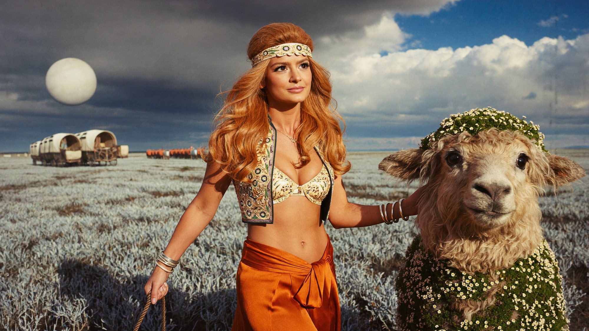 Carla as a desert trader in a beaded headband and orange sarong, walking beside a woolly alien creature, wagons behind her.