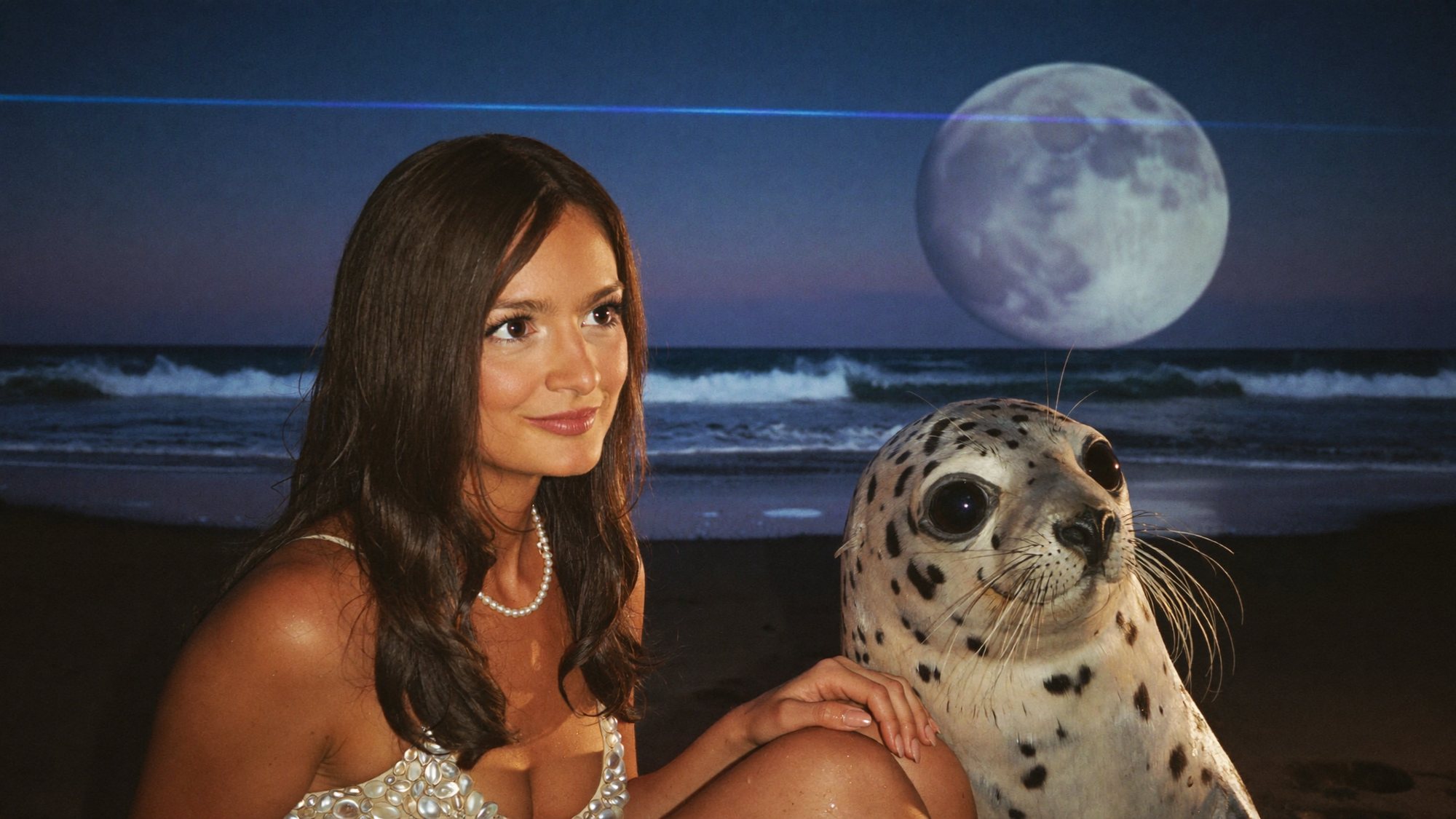 Carla in a sequinned dress sitting on a moonlit alien beach beside a friendly spotted seal-like creature.