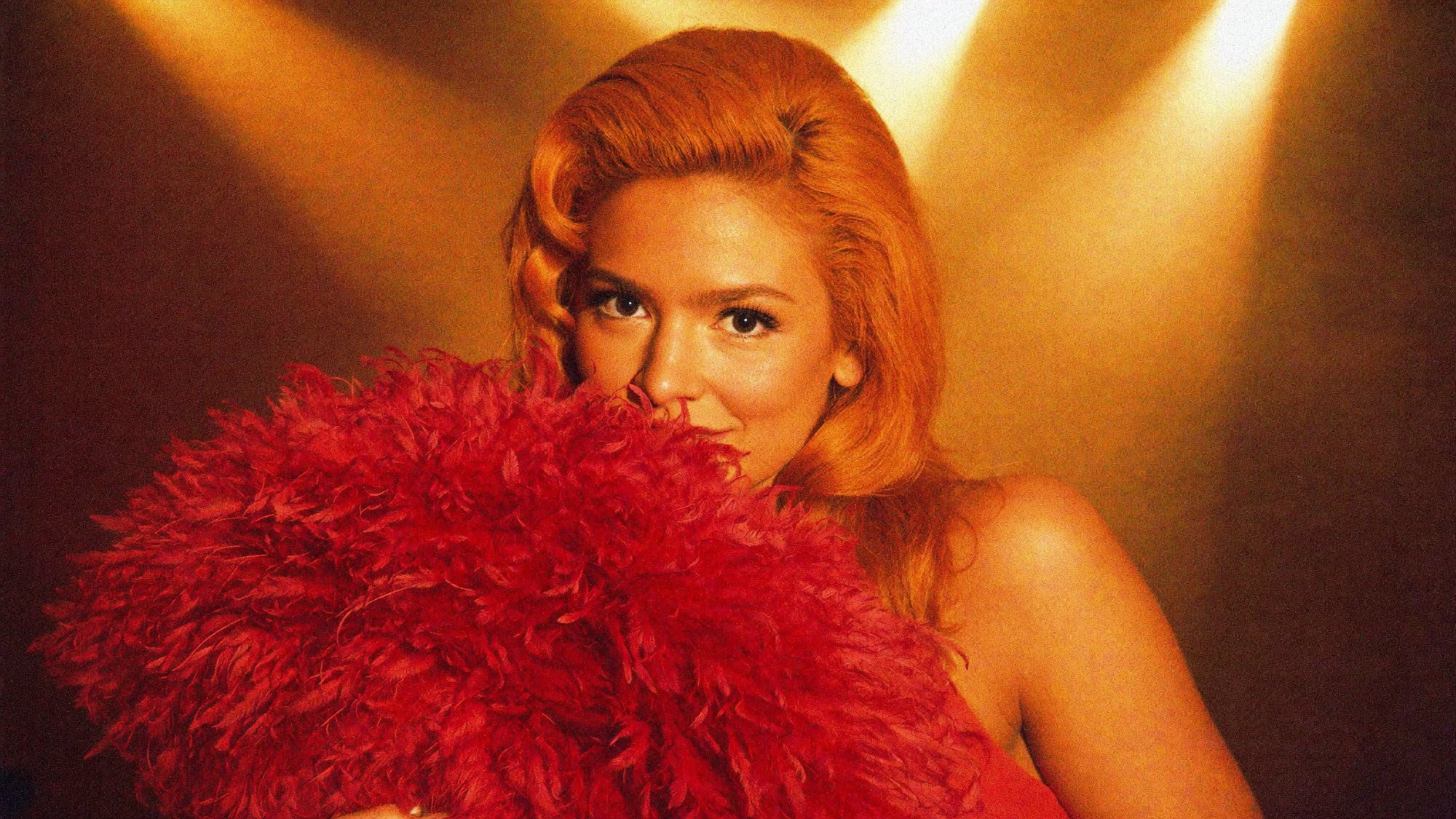 Orange-haired Carla winking over a red feather fan in amber stage light.