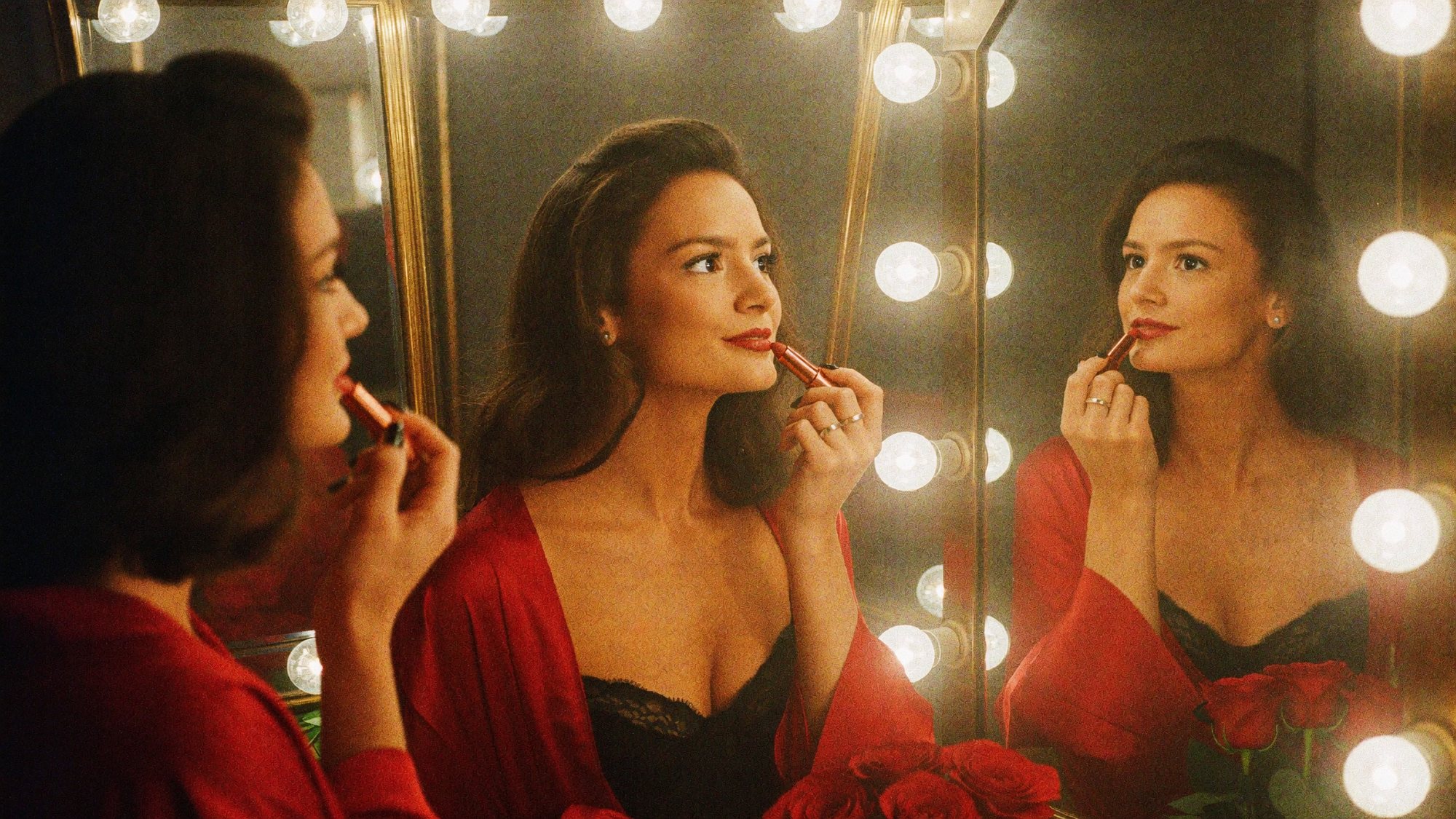 Carla in a red robe applying lipstick in a bulb-lit dressing-room mirror, reflected three times.