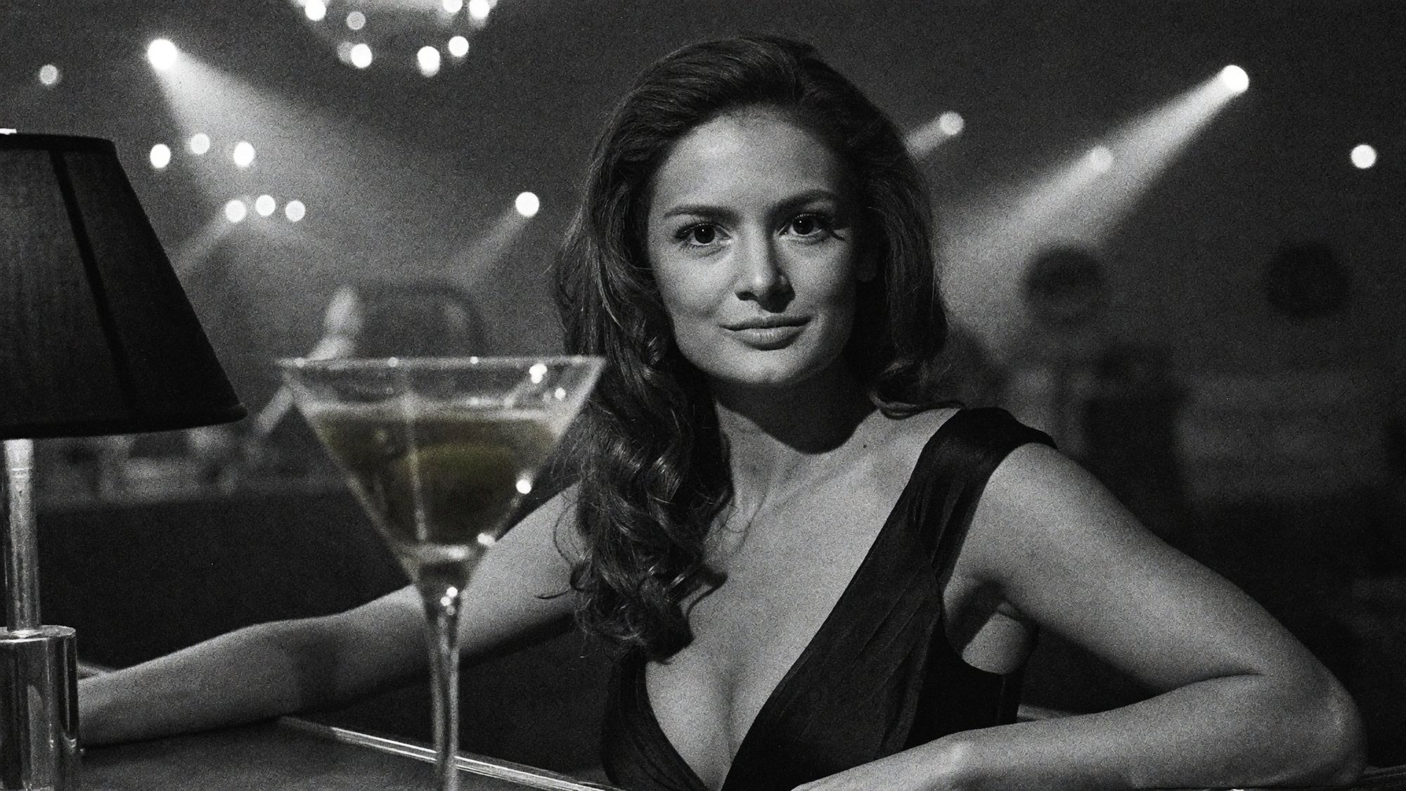 Carla in black and white leaning on a bar beside a martini glass.